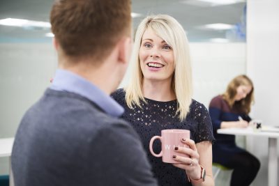 A female worker standing in an office, holding a mug and smiling whilst speaking to a male coworker in the foreground whose face is not visible