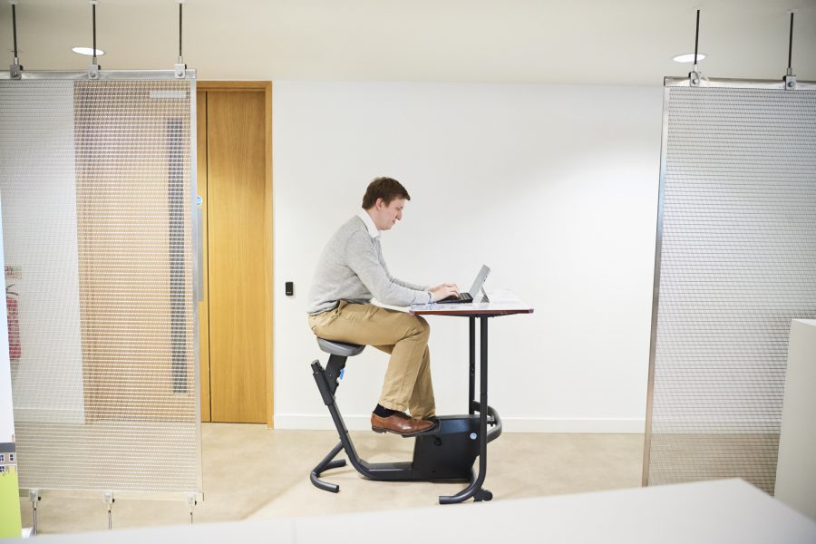 Man sitting at a pedal desk - a desk with a bicycle seat and bicycle pedals - in an office, working on a tablet computer