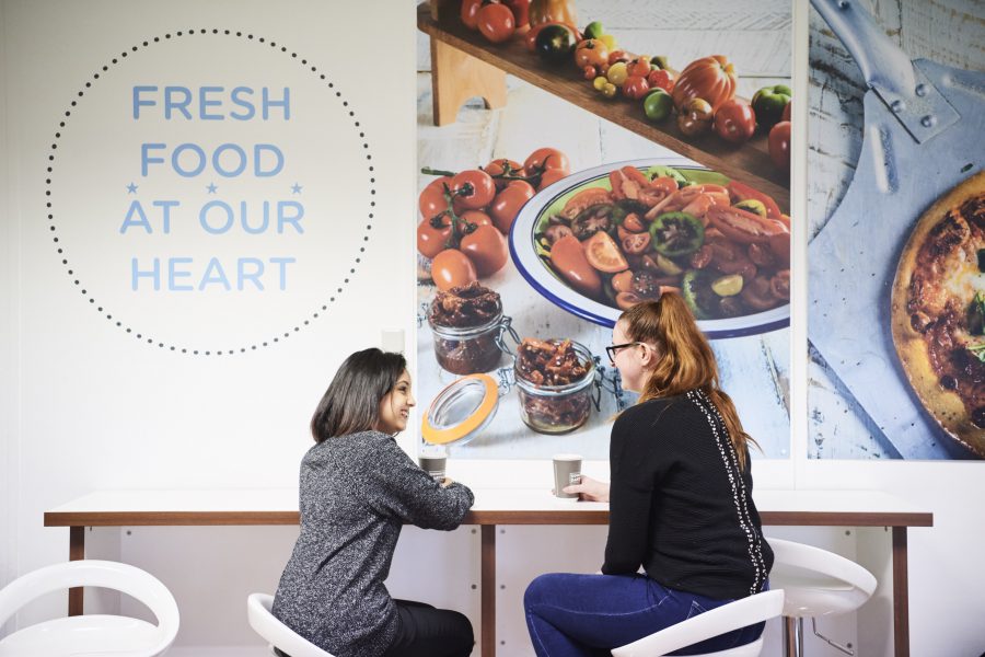 Two females sitting on chairs next to a wall in office canteen having a conversation