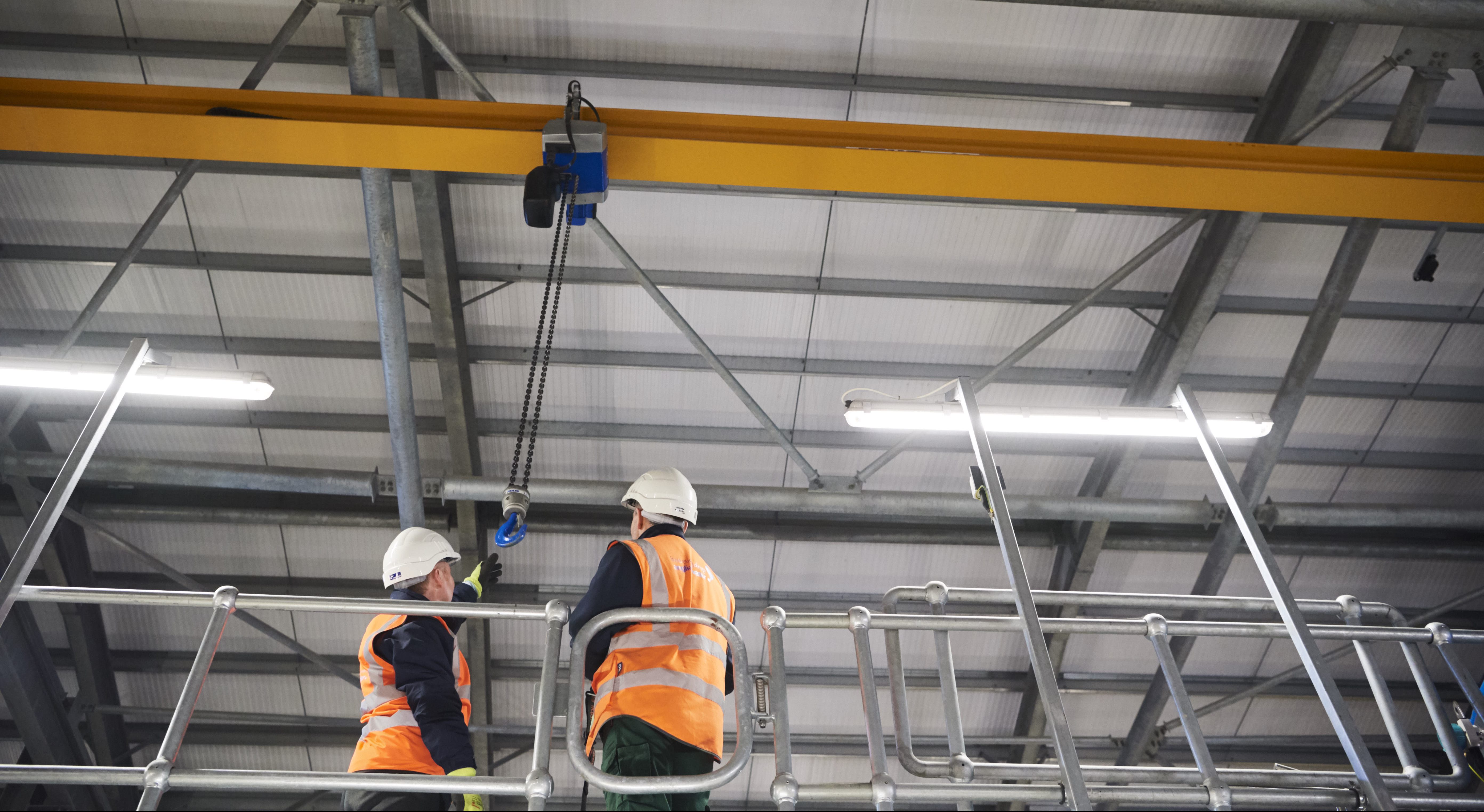 Shot from below of two males in high vis jackets and hard hats reaching from a hook on a crane