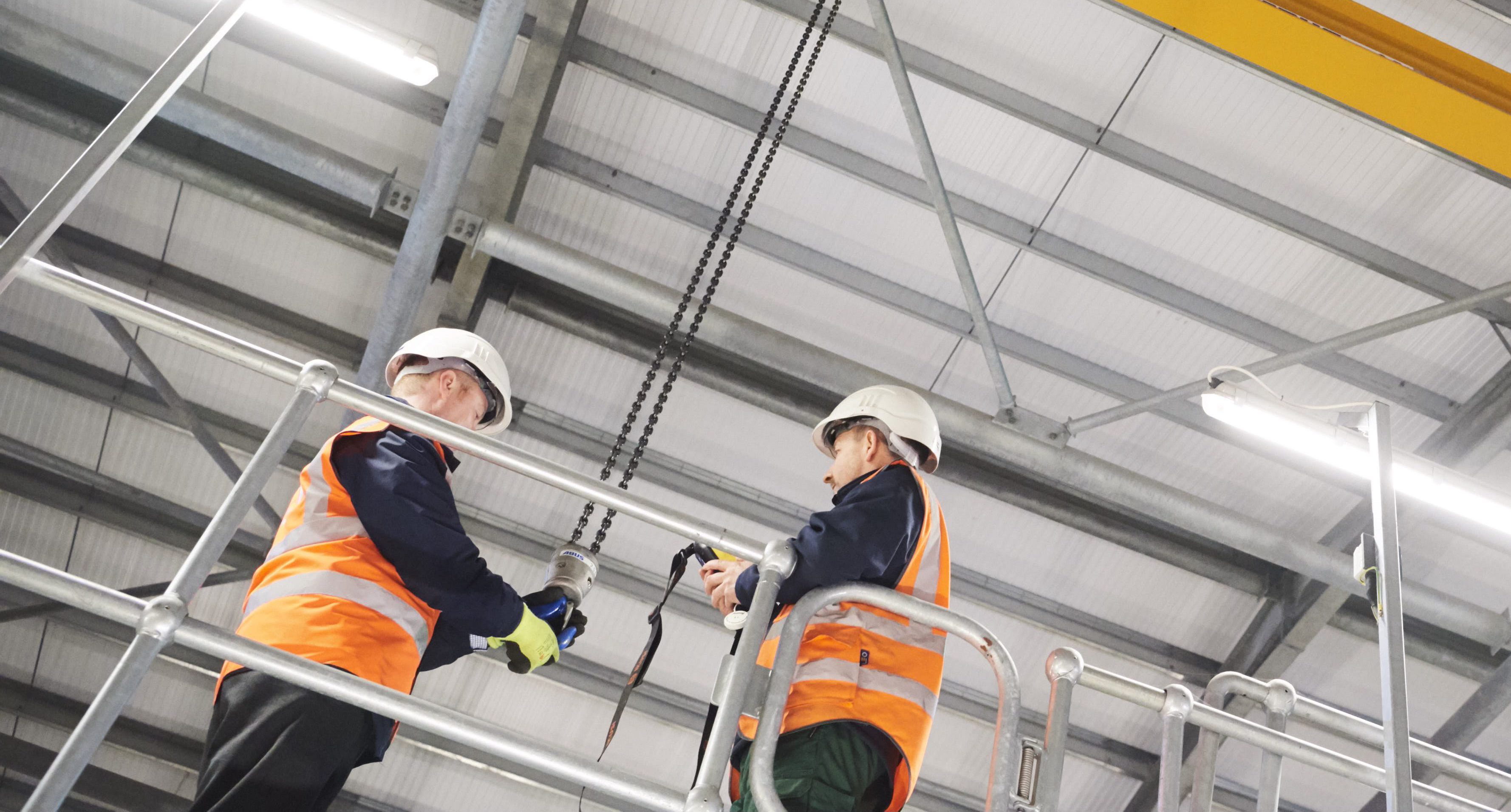 Looking up at 2 male colleagues - both in hard hats and helmets, one holding a hook on a chain dangling from the facility ceiling