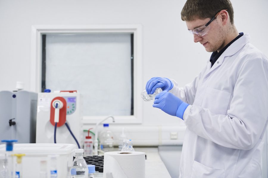 Male in laboratory environment with lab coat and gloves pouring one liquid into an unseen container