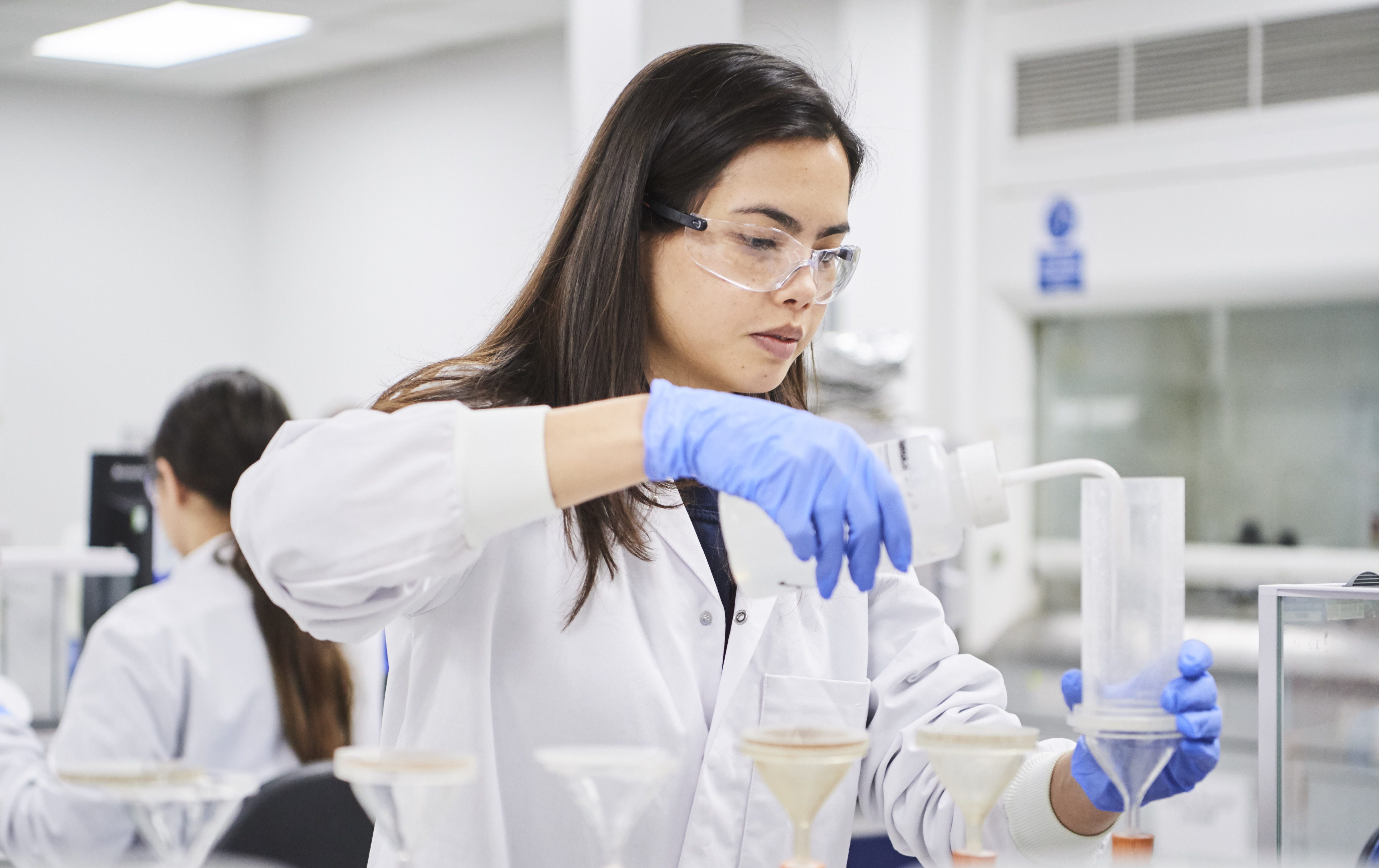 Female in lab environment in lab coat and gloves transferring liquid into a funnel