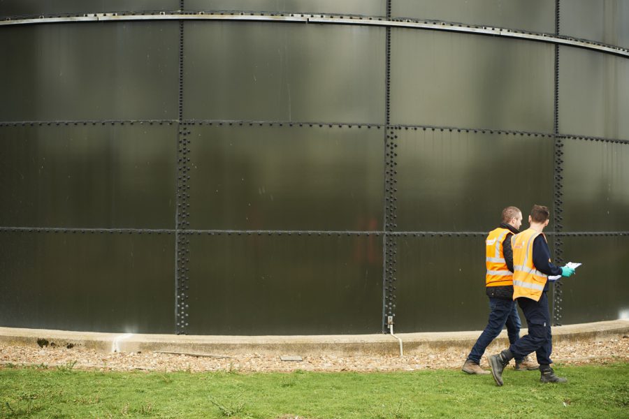 Two males in high vis jackets walking past green water cylinder