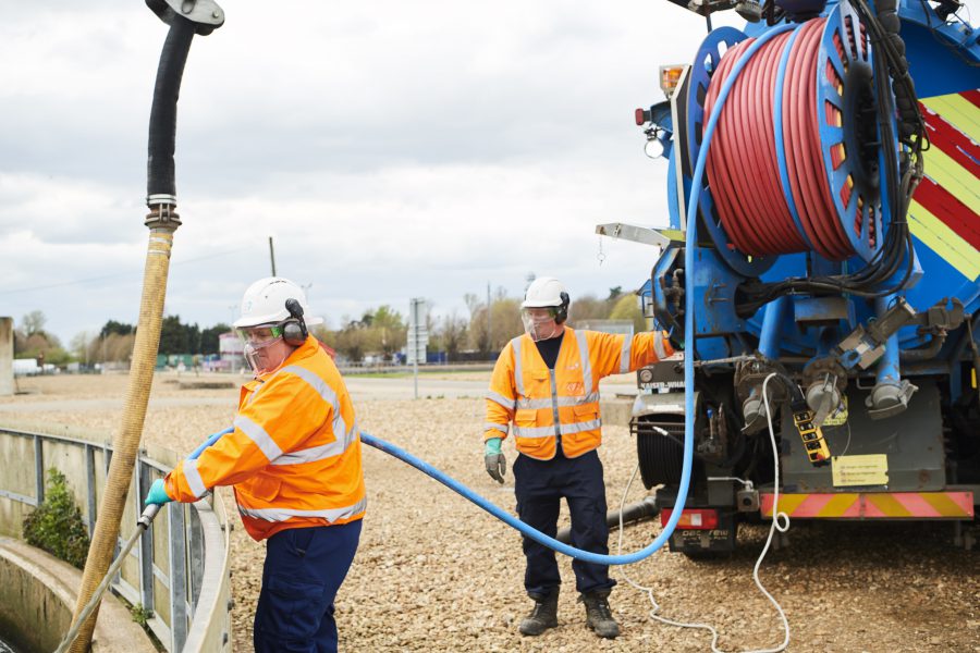 Two males in high vis jackets, hats and masks using the hose from a large tank vehicle