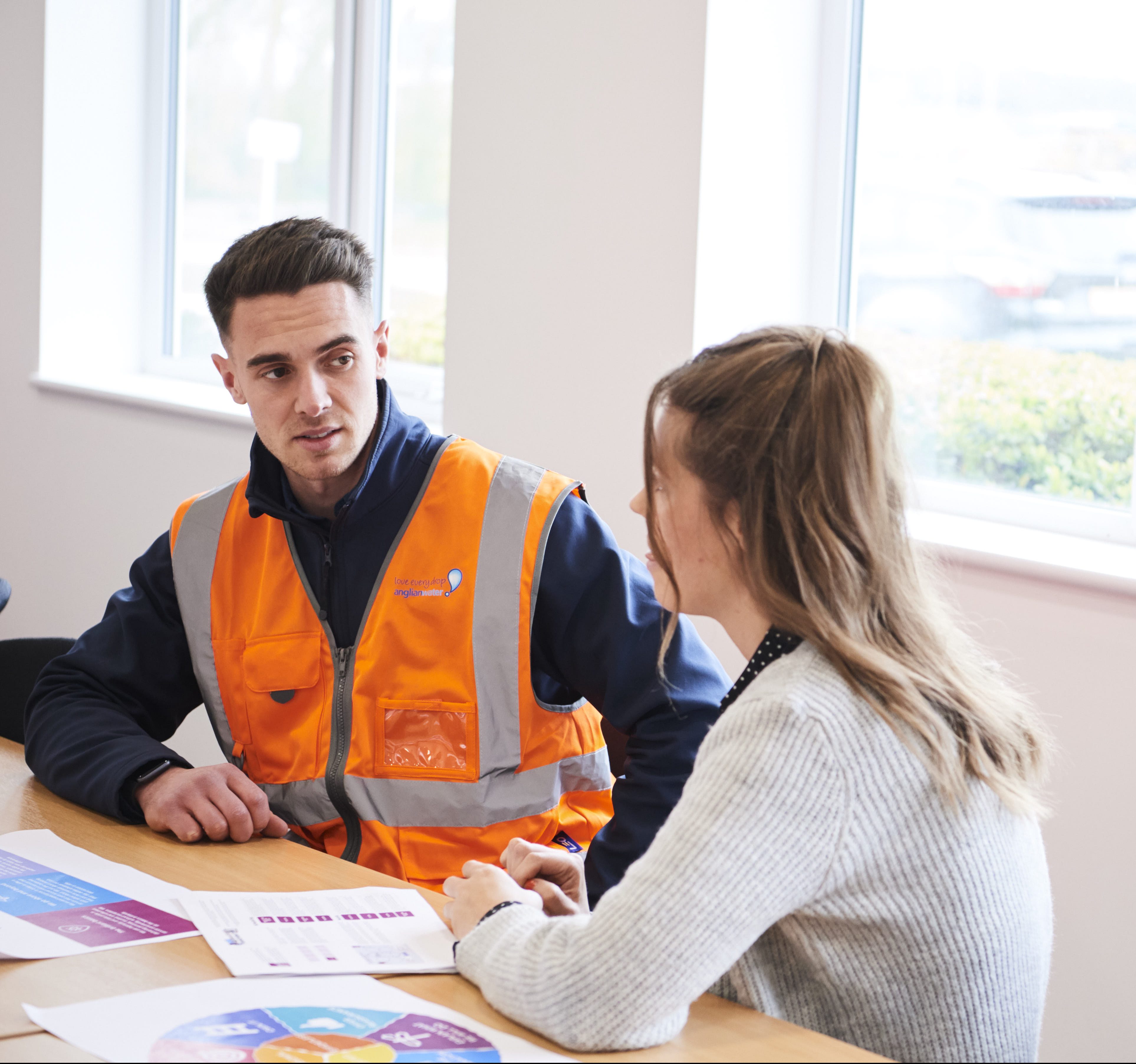 One male graduate in high vis and one female looking at papers with charts on at table in office environment