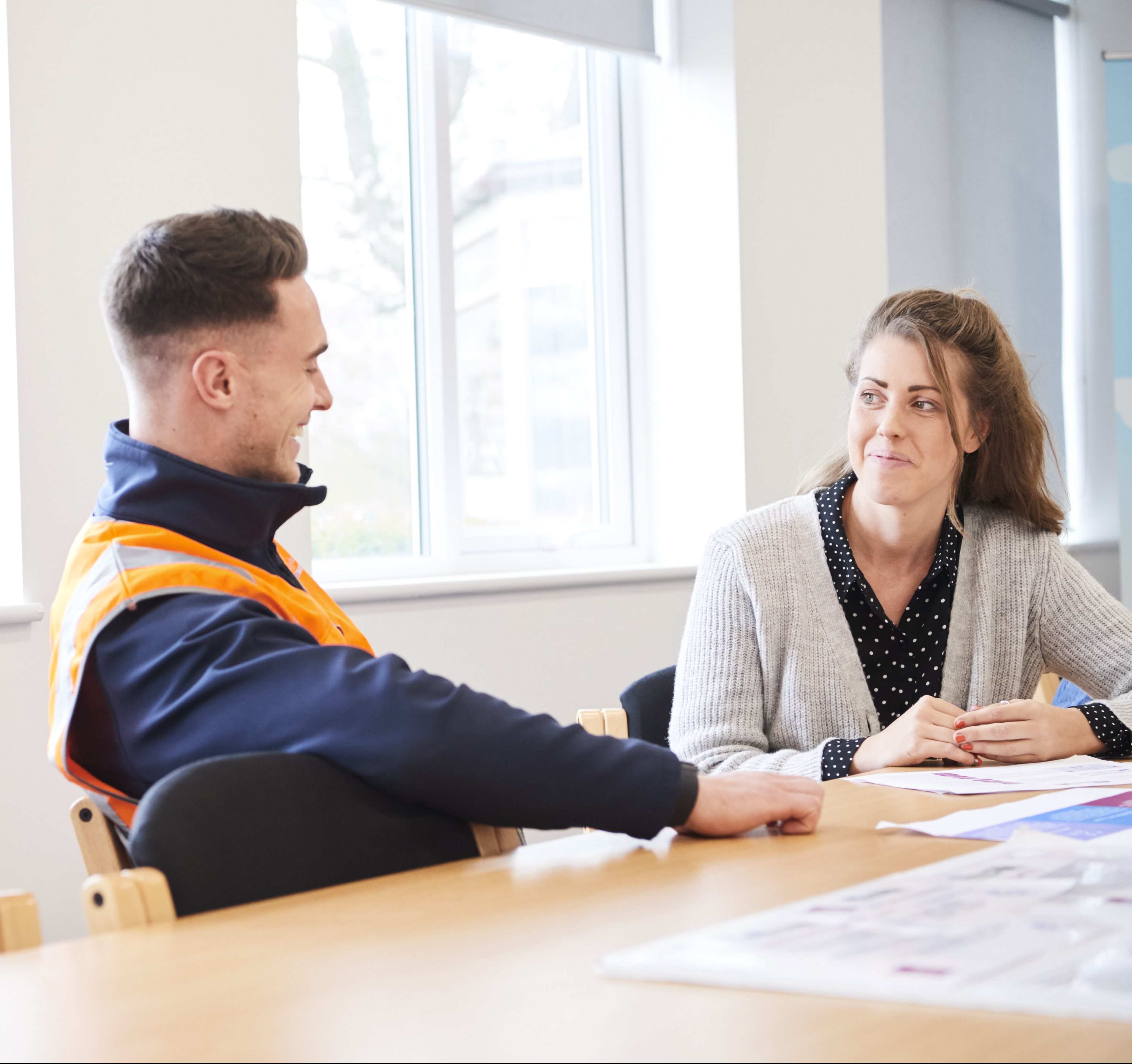 Two colleagues, a male in a high vis vest and a female in typical work clothes, sitting at a table in an office, talking